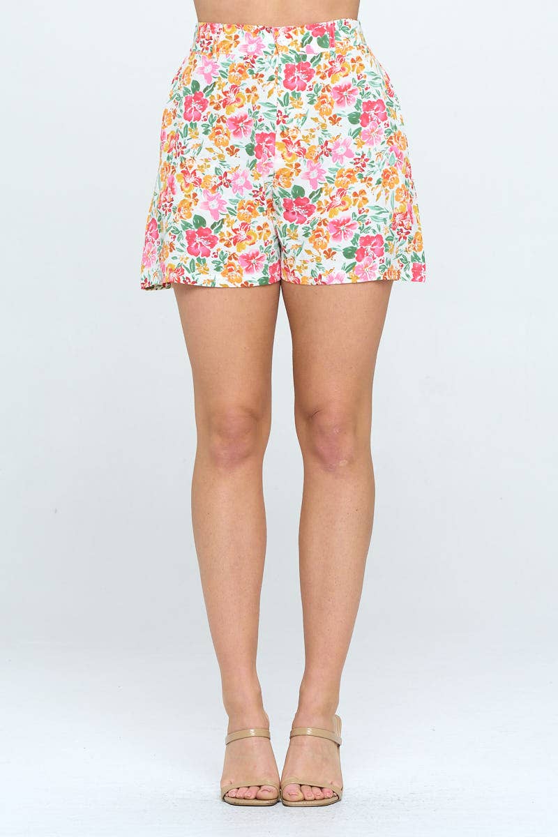 High-waisted floral shorts with a pink, orange, and green tropical print on white. Relaxed fit, mid-thigh length, styled with nude heels.