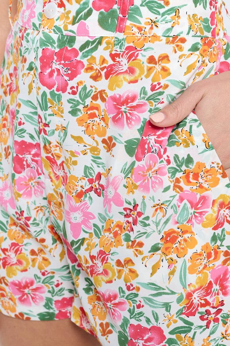 A close-up view of high-waisted floral shorts, showcasing the pink, orange, and green tropical flower print on white fabric. The model’s hand rests inside the side pocket, highlighting the pocket design and fabric texture.