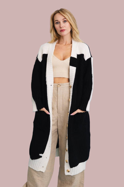 A woman wearing a color block button-up knit cardigan in black and white, with wooden buttons, paired with a beige top and beige trousers.