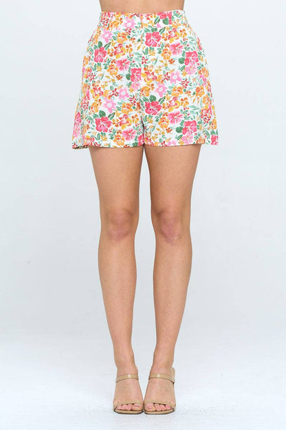 High-waisted floral shorts with a pink, orange, and green tropical print on white. Relaxed fit, mid-thigh length, styled with nude heels.